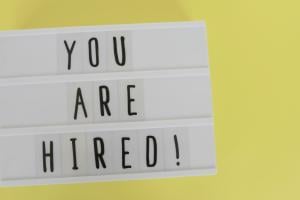 Against a yellow background reader board of black text that says, "YOU ARE HIRED!"