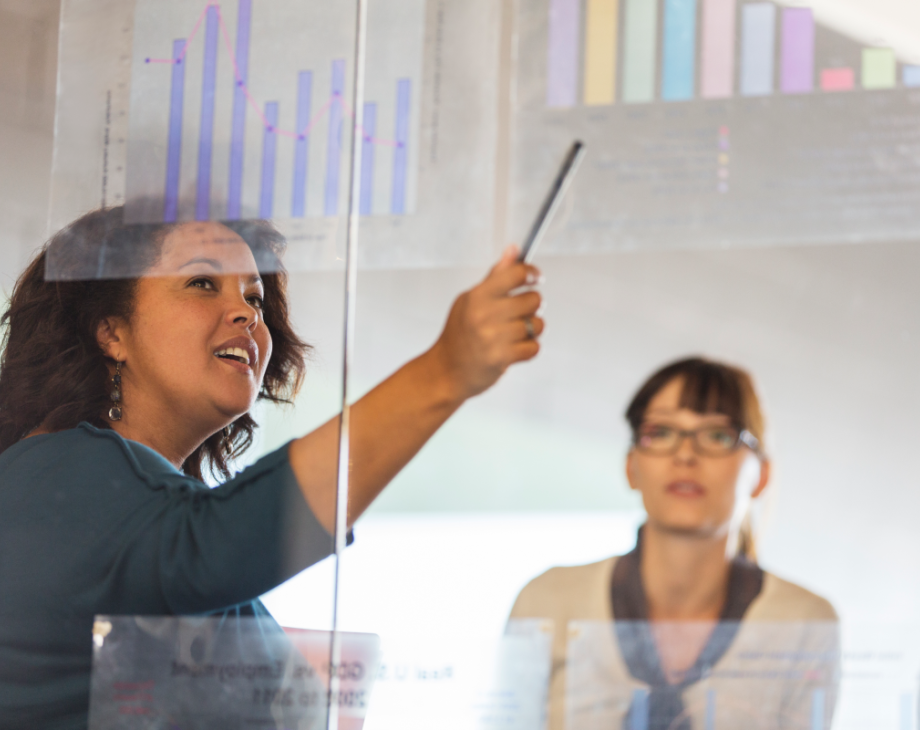 Color photo of woman of color pointing to graphs in front of two white colleagues