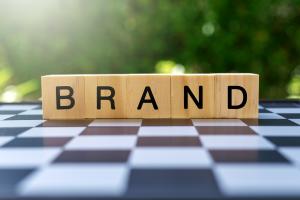 Stock image of Scrabble tiles spelling "brand"sit atop a black-and-white checkerboard with greenery in the background 