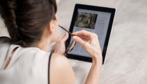 White woman holds glasses in right hand while looking at a digital tablet
