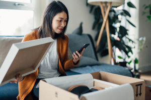 Young Asian woman on couch looks at her mobile phone as she opens a package