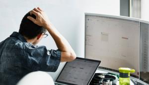 White, bespectacled man scratches his head in front of a laptop screen and computer monitor
