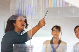 Color photo of woman of color pointing to graphs in front of two white colleagues