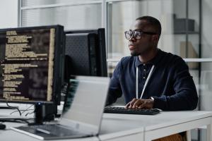 Color photo of young Black man sitting in front of a computer monitor at an office 