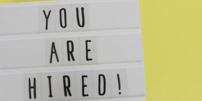 Against a yellow background reader board of black text that says, "YOU ARE HIRED!"
