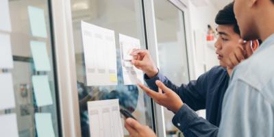 Color photo of two men drawing on a glass wall in a company office setting