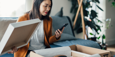 Young Asian woman on couch looks at her mobile phone as she opens a package