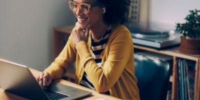 Bespectacled black woman in yellow cardigan smiles at her open laptop screen