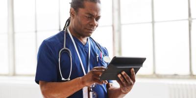 Black male doctor with stethoscope and in blue scrubs looks at tablet