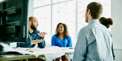 Color photo of four people sitting at a table in front of a computer monitor in an office