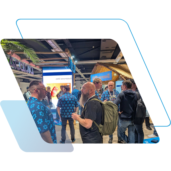 A group of people network at a tropical-themed trade show booth. Several men wear matching blue floral shirts, while a man in the foreground with a long beard and olive-green backpack talks with them near a "tiki" style structure and a digital display.