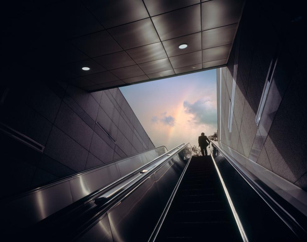 Man riding up an escalator toward rainbow in sky