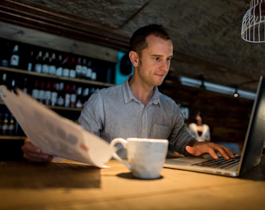 Man working in cafe on laptop with coffee mug and papers