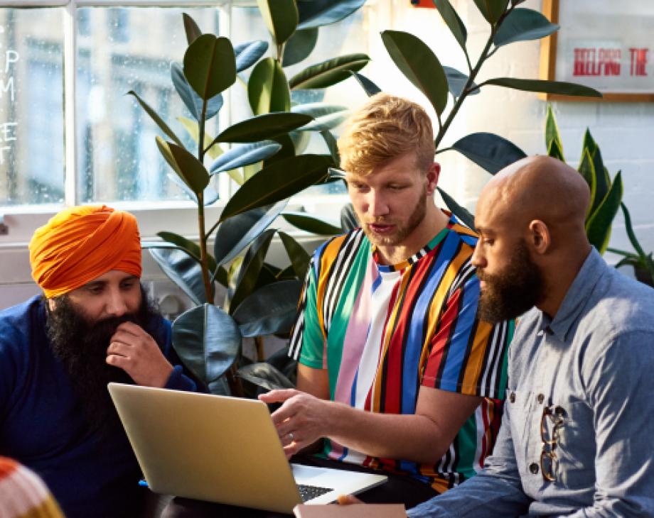 3 men of various races sit in brightly colored clothes and look at a laptop screen