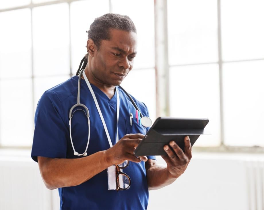 Black male doctor with stethoscope and in blue scrubs looks at tablet