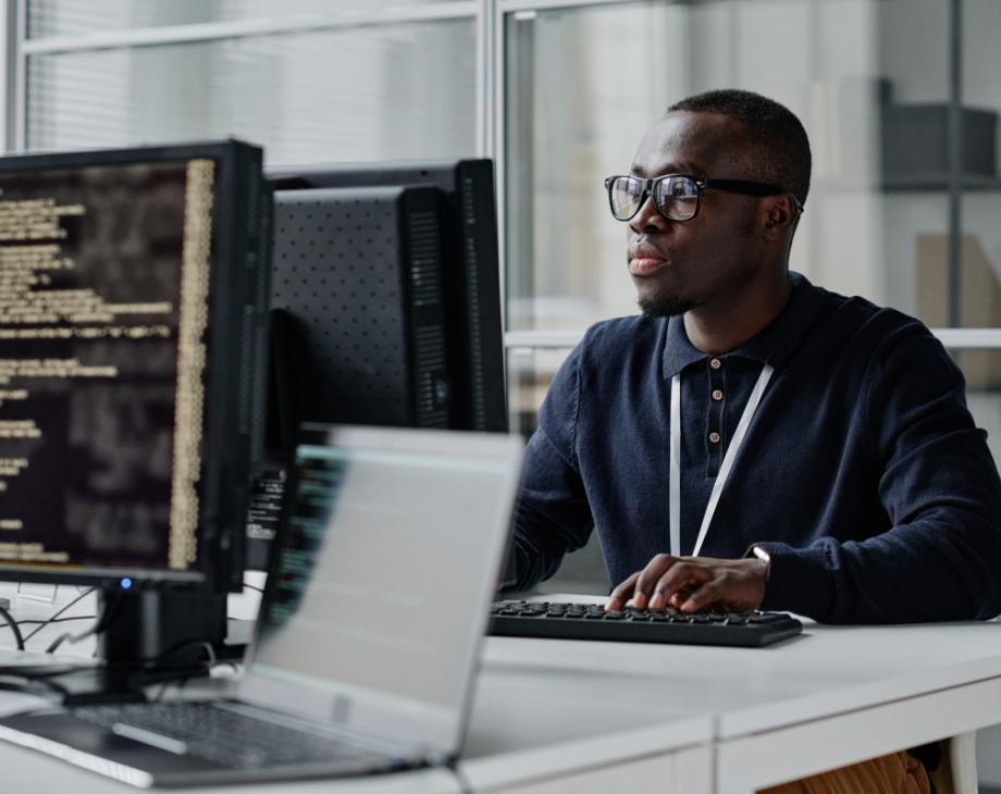Color photo of young Black man sitting in front of a computer monitor at an office 