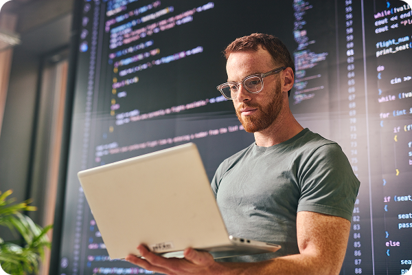 Man in glasses holding laptop in front of digital code display screens